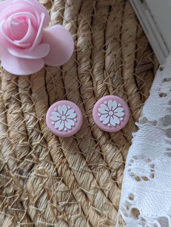 two pink flower-shaped earrings with white flower designs, placed on a woven surface next to a white lace fabric.