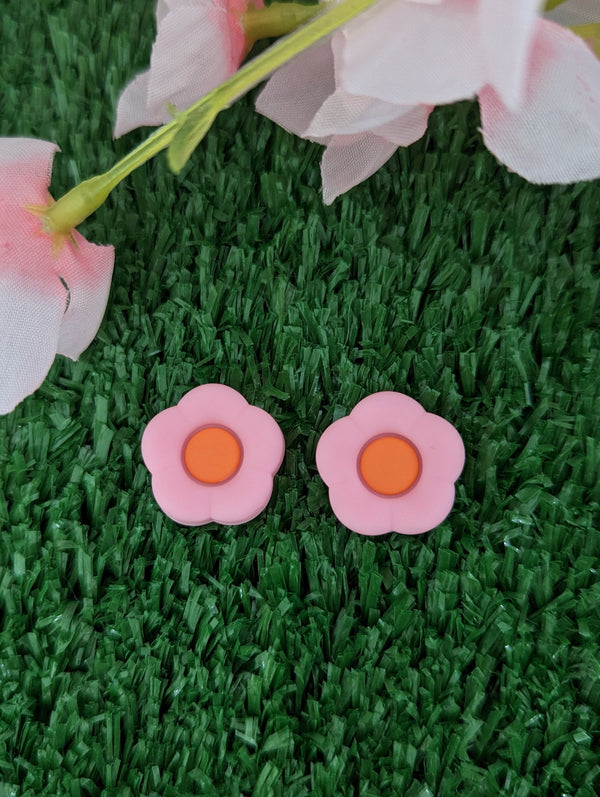 Two pink flower-shaped earrings are placed on a green grass surface, with a pink flower-like object in the background.