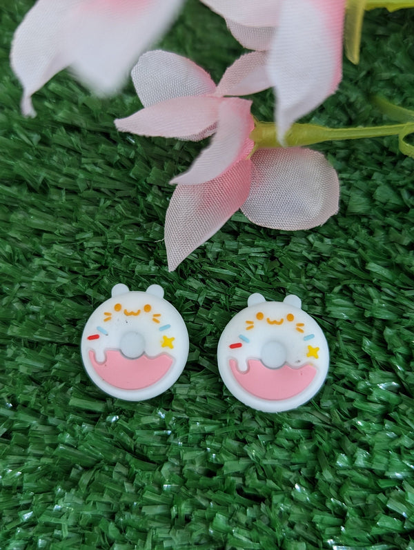 two white donut-shaped earrings with pink and yellow accents, placed on a green surface with pink and white flowers in the background.