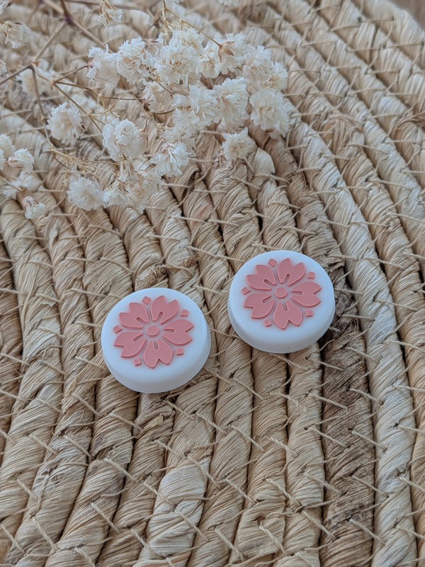 two white circular objects with pink flower designs on them, placed on a woven surface.