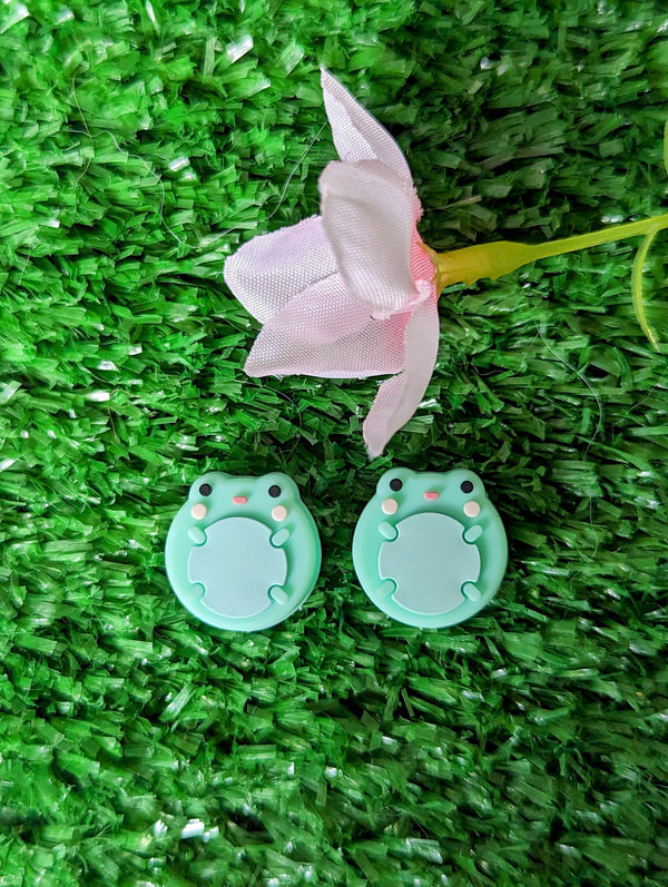 two green frog-shaped objects and a pink flower on a green grassy surface.