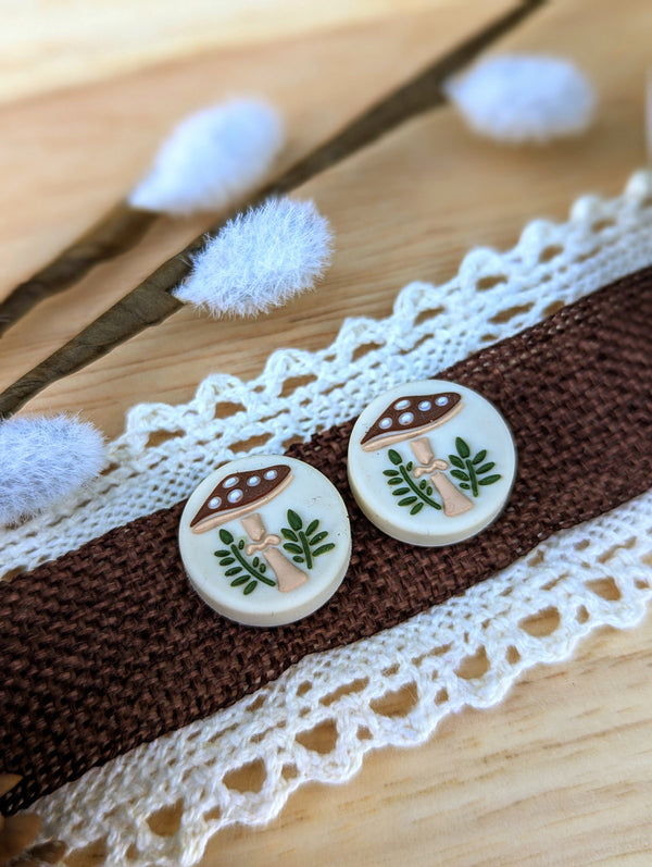 two circular buttons with mushroom and leaf designs, placed on a brown fabric with a lace-like pattern.