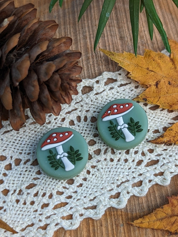 two green buttons with mushroom designs, placed on a white lace doily. The buttons are surrounded by pine cones and autumn leaves, creating a natural and rustic setting.