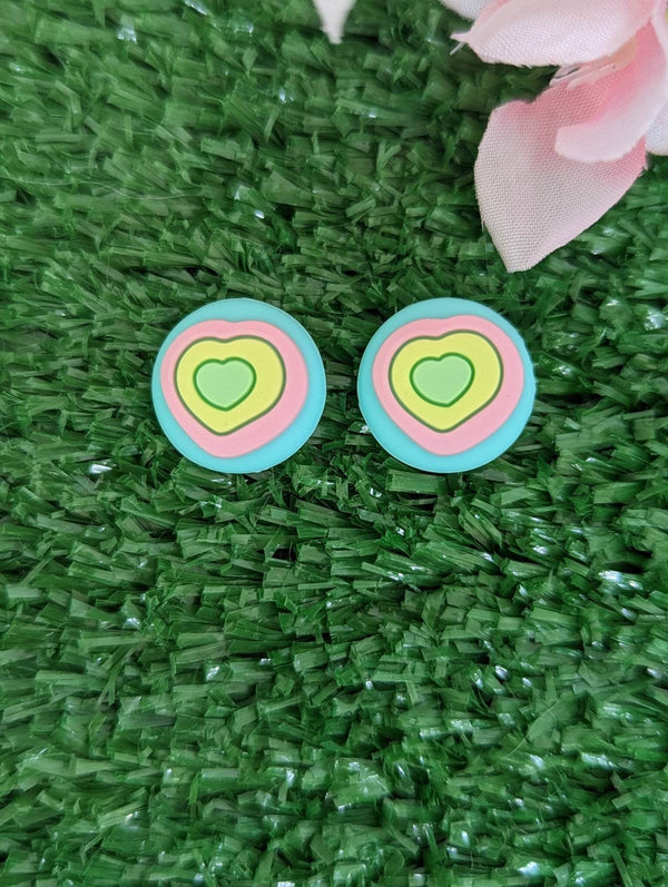 Two heart-shaped earrings with colorful, pastel-colored designs are placed on a green surface, with a pink flower petal visible in the background.