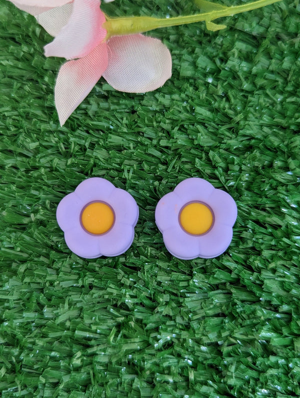 two purple flower-shaped earrings with yellow centers, placed on a green grassy surface.