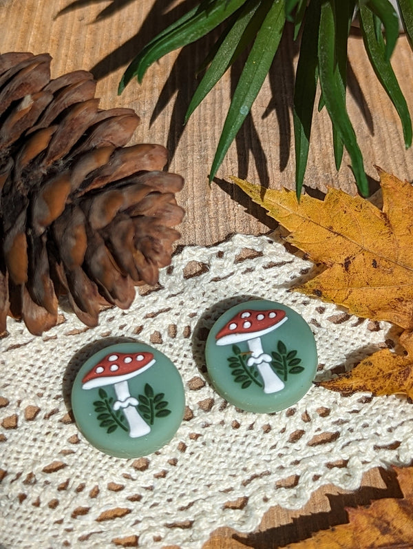 two green mushroom-shaped buttons with white spots, placed on a white lace doily. The buttons are surrounded by autumn leaves, pine cones, and a lace doily, creating a rustic and natural setting.